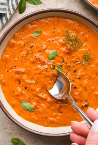 A large ceramic bowl of thick and creamy tomato and red pepper soup, garnished with fennel fronds and fresh basil leaves. A spoon is dipping into the soup to show the luscious texture.