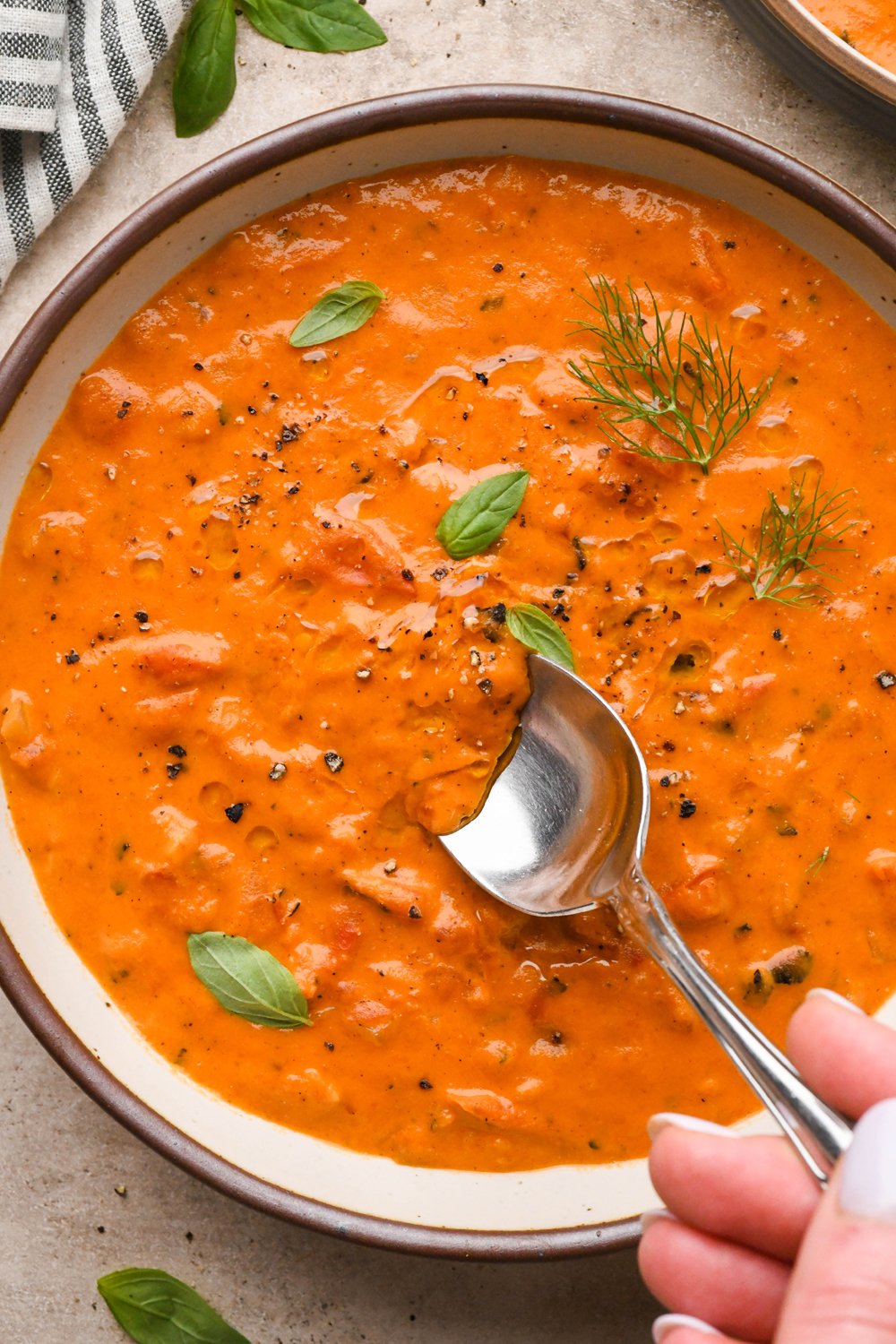 A large ceramic bowl of thick and creamy tomato and red pepper soup, garnished with fennel fronds and fresh basil leaves. A spoon is dipping into the soup to show the luscious texture.