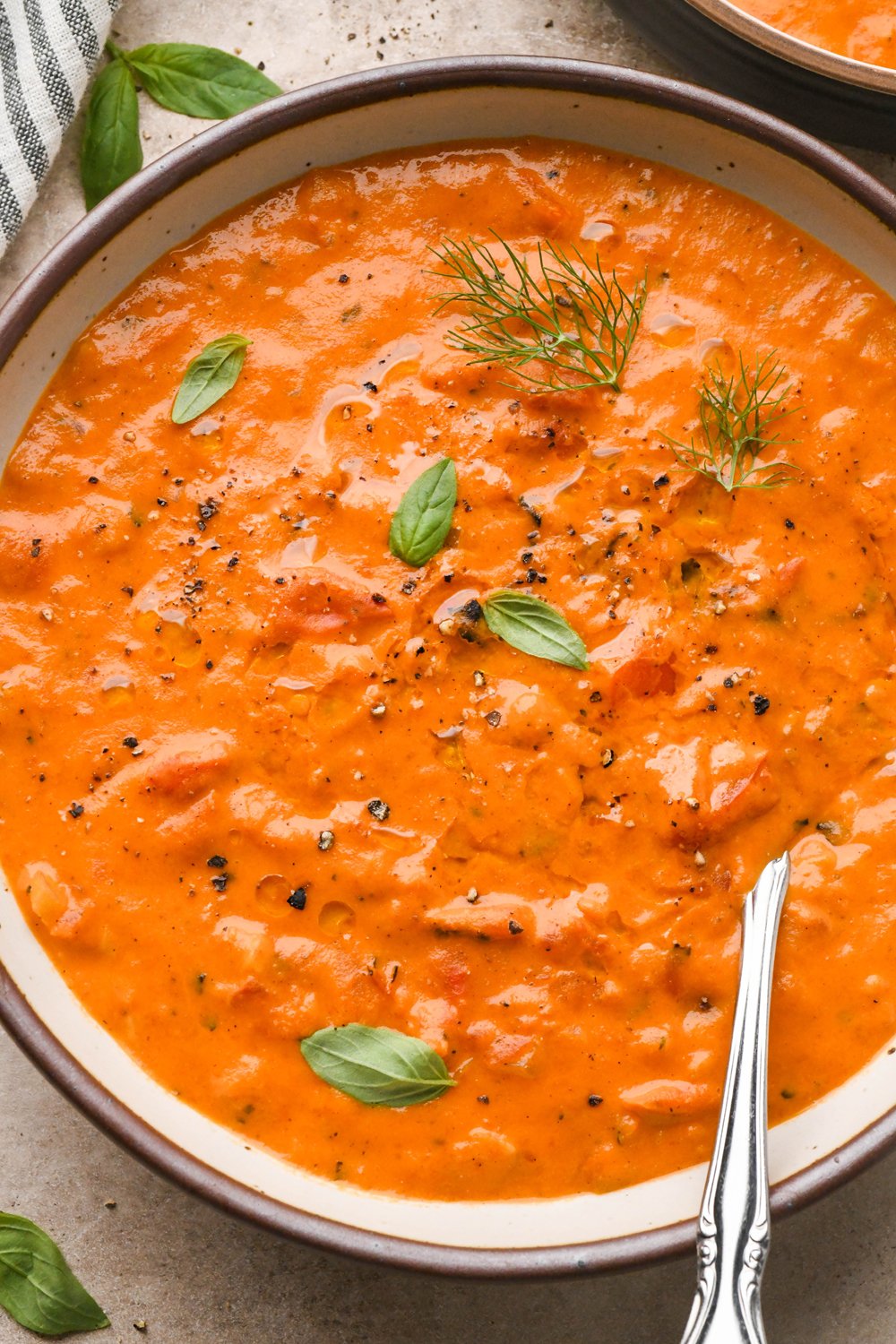 A large ceramic bowl of thick and creamy tomato and red pepper soup, garnished with fennel fronds and fresh basil leaves. A spoon is angled into the bowl to show the luscious texture.