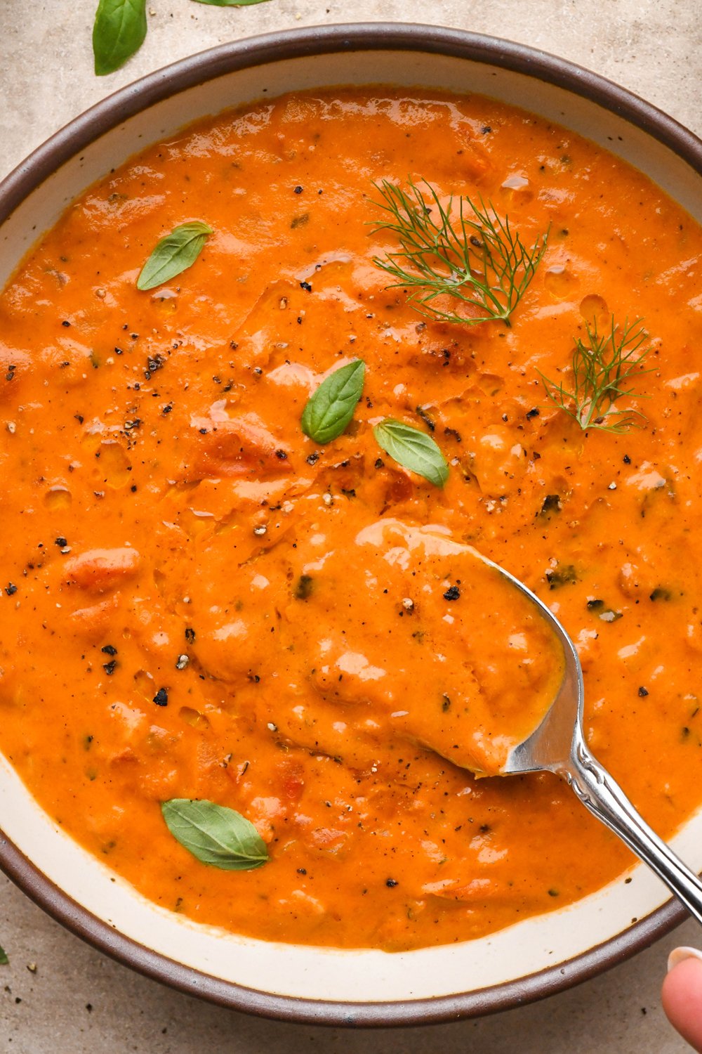 A large ceramic bowl of thick and creamy tomato and red pepper soup, garnished with fennel fronds and fresh basil leaves. A spoon is dipping into the soup to show the thick and creamy texture.