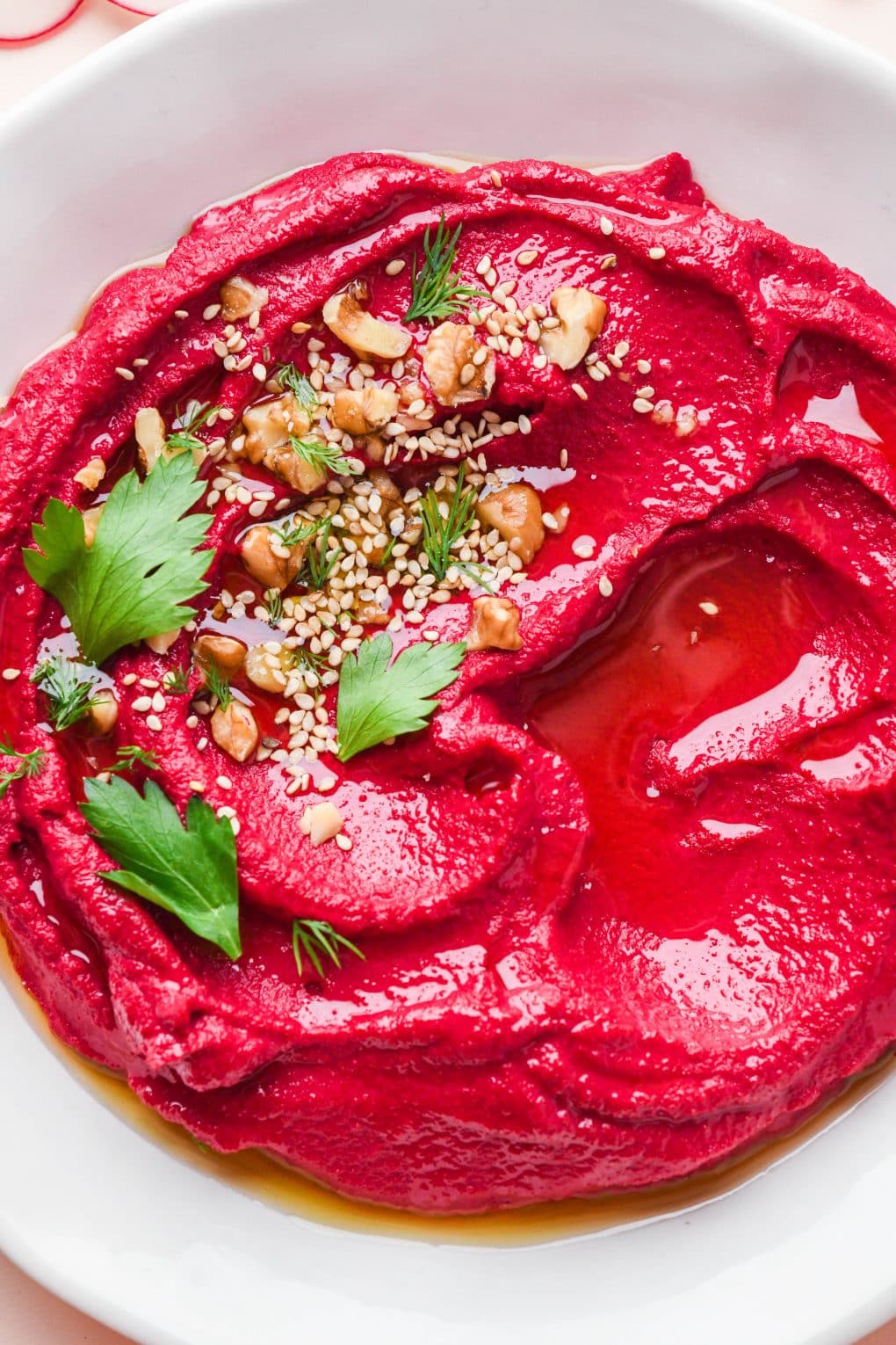 Bright red cashew beet dip in a shallow bowl topped with nuts, seeds, and some fresh herbs.