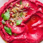 Bright red cashew beet dip in a shallow bowl topped with nuts, seeds, and some fresh herbs.