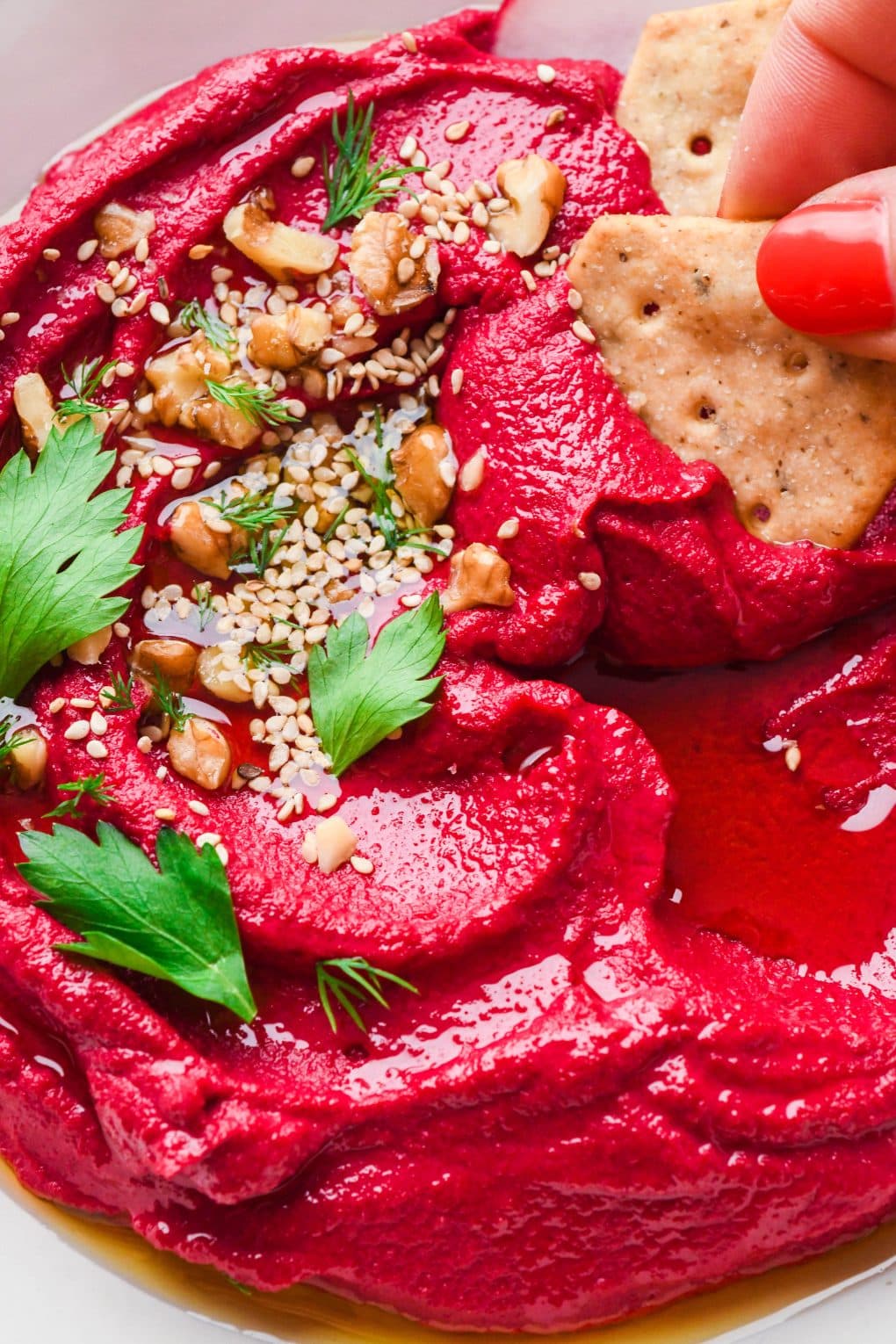 Bright red cashew beet dip in a shallow bowl topped with nuts, seeds, and some fresh herbs with a hand dipping a cracker into the creamy dip.