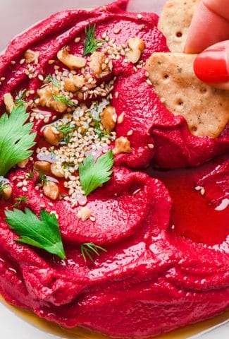 Bright red cashew beet dip in a shallow bowl topped with nuts, seeds, and some fresh herbs with a hand dipping a cracker into the creamy dip.