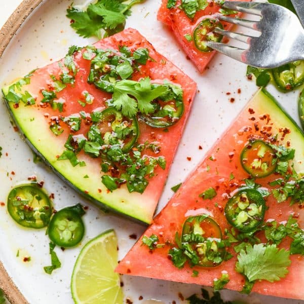 Spicy watermelon salad wedges on a ceramic plate topped with cilantro, jalapeño, lime dressing with a fork digging into a bite of the watermelon.