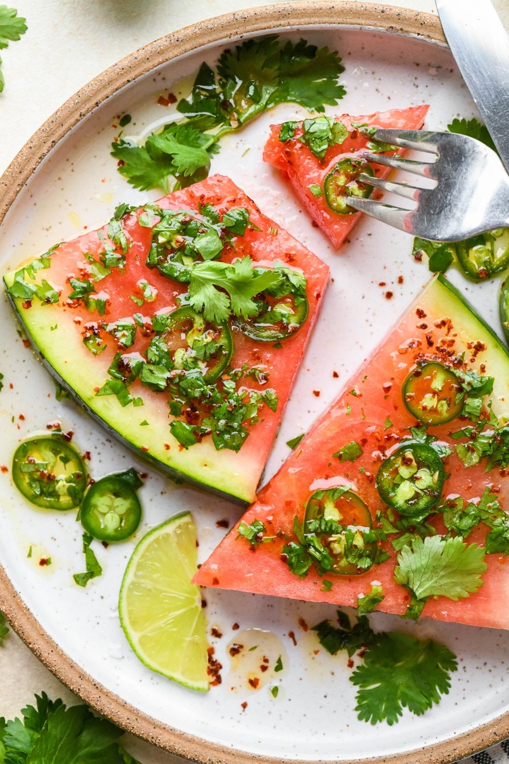 Spicy watermelon salad wedges on a ceramic plate topped with cilantro, jalapeño, lime dressing with a fork digging into a bite of the watermelon.