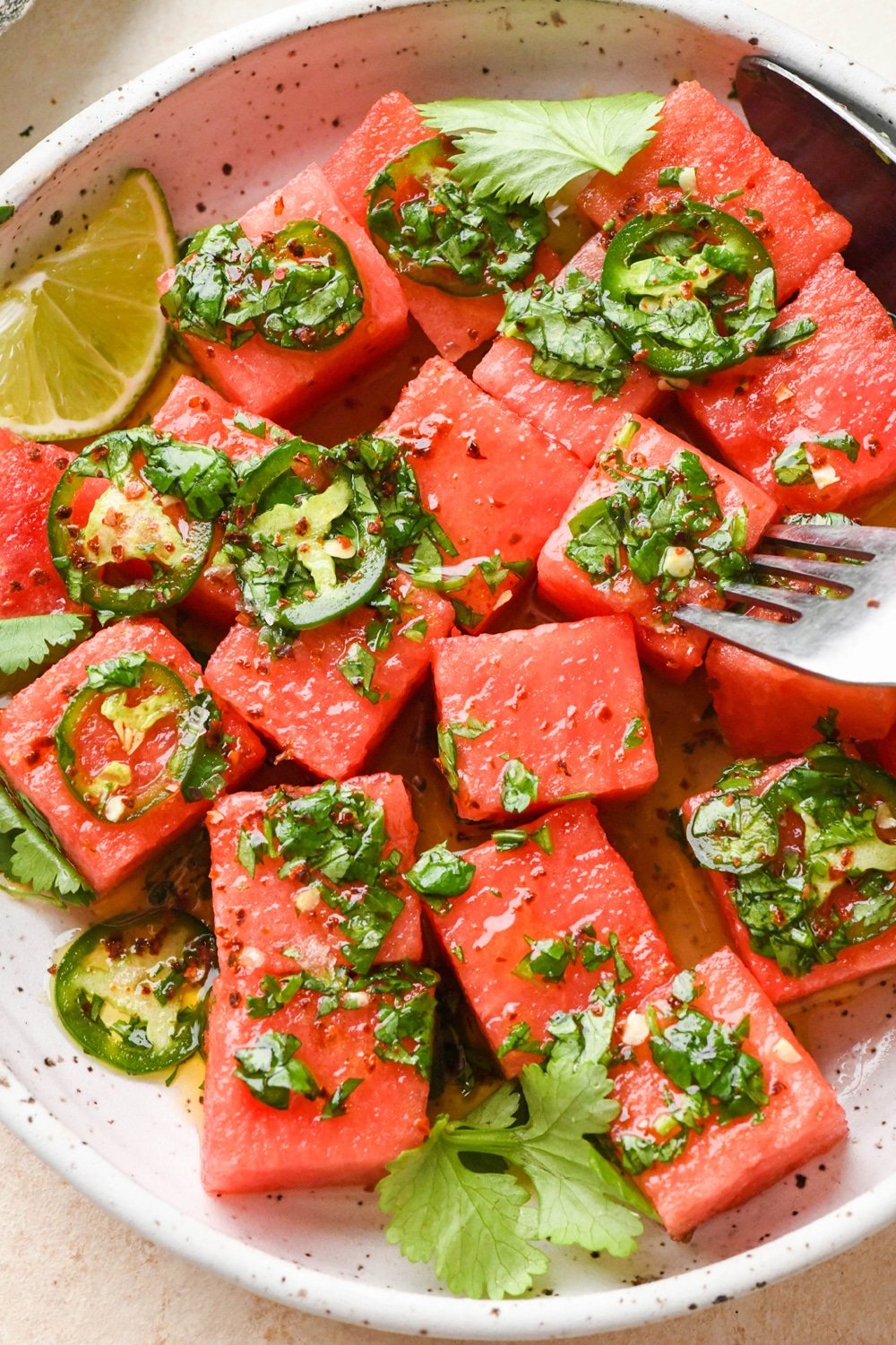 How to make spicy watermelon salad: Cubed watermelon in a bowl with dressing over the top and a fork piercing a piece of watermelon.
