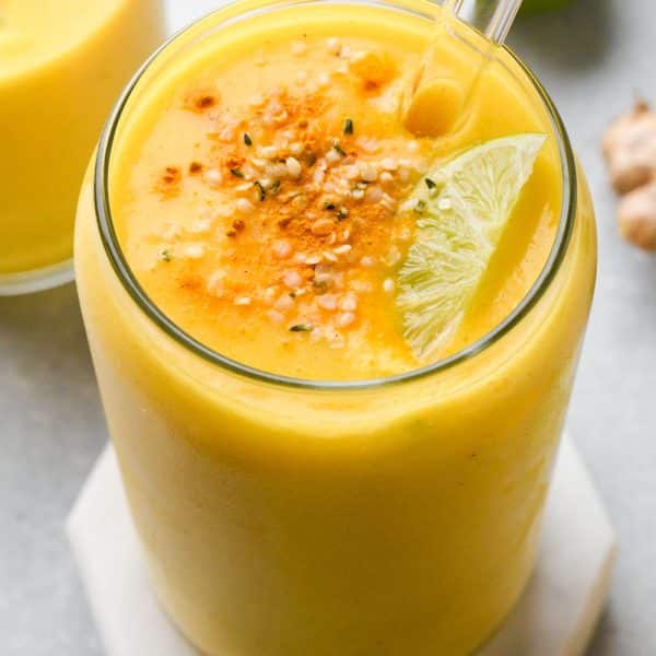 A cup tall filled with a vibrant yellow mango ginger turmeric smoothie, garnished with a sprinkle of dried turmeric, hemp seeds, and a lime wedge. On a light blue background, and surrounded by fresh ginger, lime, and mango.
