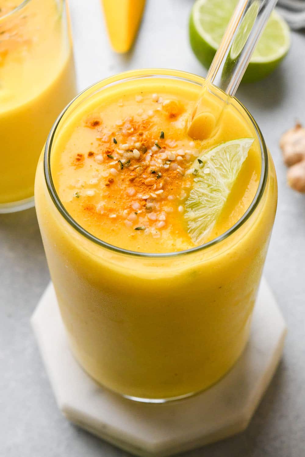 A cup tall filled with a vibrant orange mango ginger turmeric smoothie, garnished with a sprinkle of dried turmeric, hemp seeds, and a lime wedge. On a light blue background, and surrounded by fresh ginger, lime, and mango.
