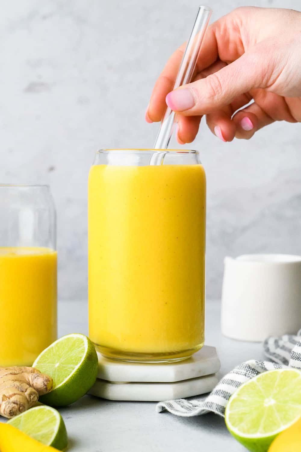 A hand placing a glass straw in a glass cup filled with vibrant yellow mango turmeric smoothie.