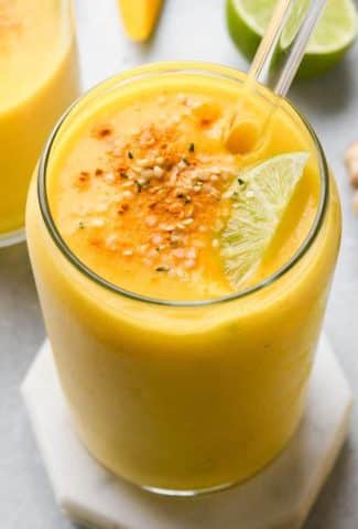 A cup tall filled with a vibrant yellow mango ginger turmeric smoothie, garnished with a sprinkle of dried turmeric, hemp seeds, and a lime wedge. On a light blue background, and surrounded by fresh ginger, lime, and mango.