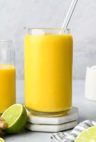 Straight on image of a tall glass cup filled with mango turmeric smoothie with a glass straw, next to a cut lime and another glass filled halfway with smoothie.