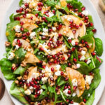 Asian pear salad with tender salad greens on a large, white, oval platter. The salad is topped with chopped toasted walnuts, pomegranate seeds, and feta cheese, and drizzled with the dressing.