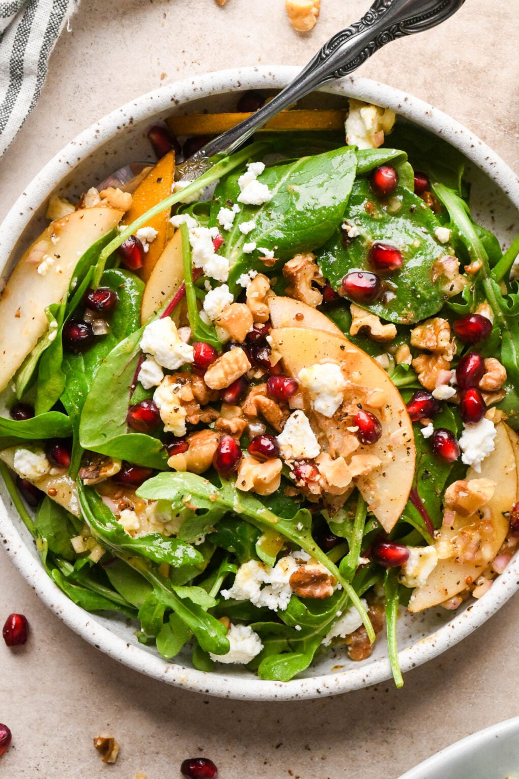 Asian pear salad in a small, white speckled serving bowl, with pomegranate, chopped walnuts, and crumbled feta cheese. The salad is dressed with a maple mustard vinaigrette, and a fork is angled into the bowl.