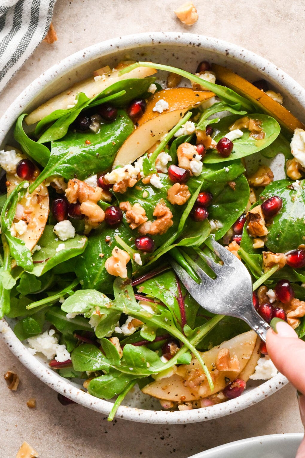 Asian pear salad in a small, white speckled serving bowl, with pomegranate, chopped walnuts, and crumbled feta cheese. The salad is dressed with a maple mustard vinaigrette, and a fork is digging into the bowl for a bite.