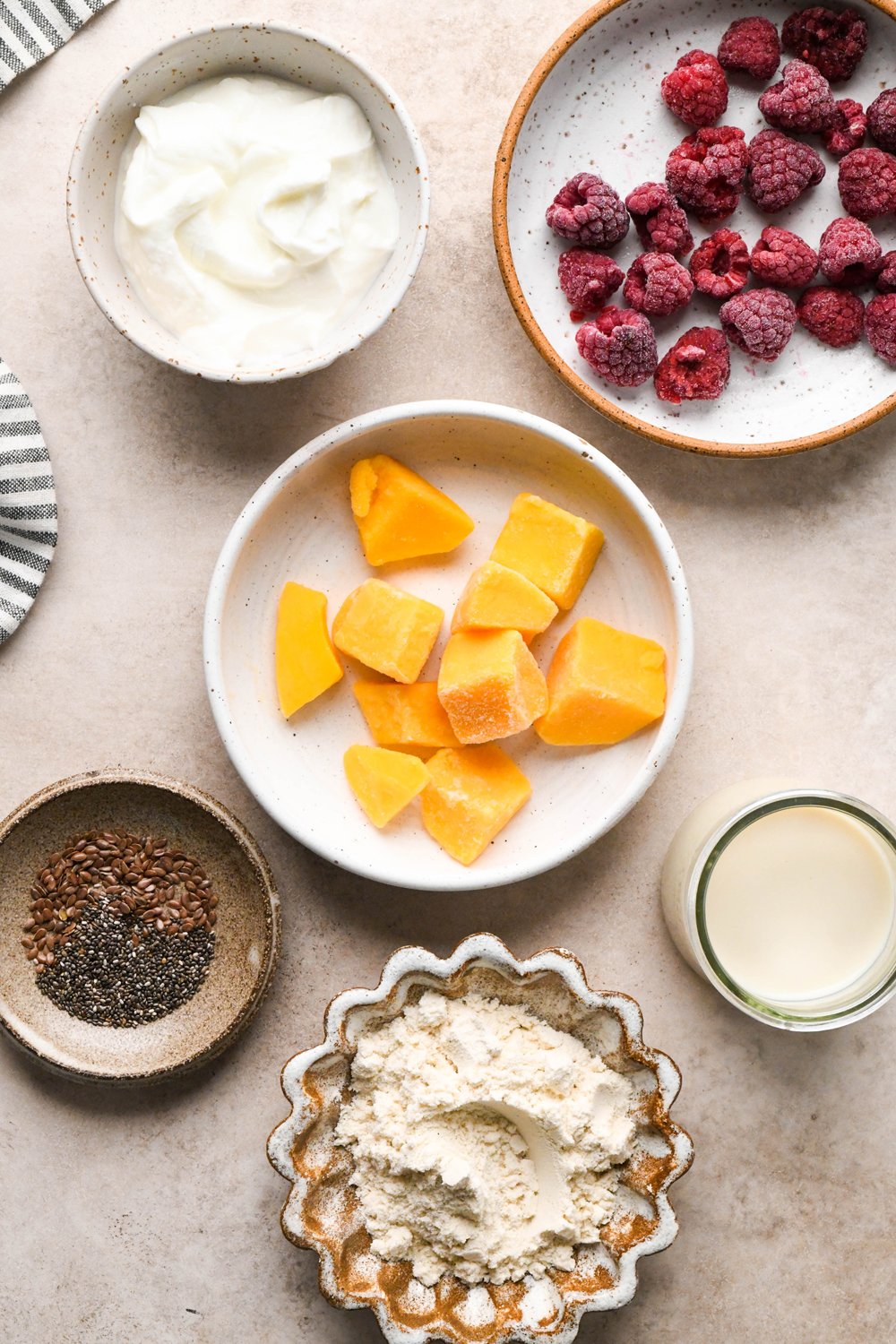 Ingredients for Raspberry Mango Smoothie with Greek Yogurt in various ceramics on a light brown creamy background.