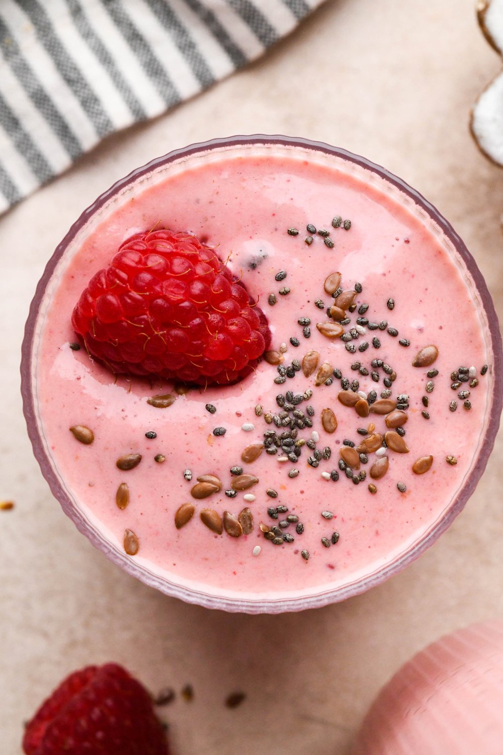 A short glass of bright pink raspberry mango smoothie garnished with a single fresh raspberry and a few chia seeds and flax seeds.