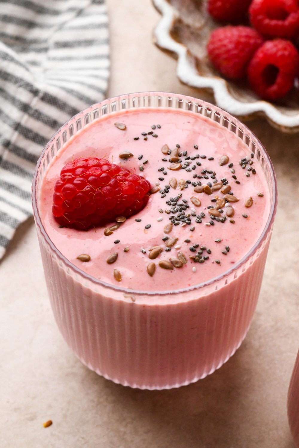 A short glass of bright pink raspberry mango smoothie topped with a single fresh raspberry and a scattering of chia seeds and flax seeds. Glass is next to a striped linen napkin and a small dish of fresh raspberries.