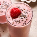 A tall glass of bright pink raspberry mango smoothie next to a smaller glass of smoothie. Both are topped with a single fresh raspberry and a scattering of chia seeds and flax seeds.