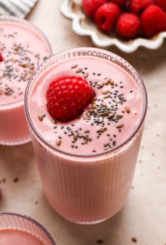 A tall glass of bright pink raspberry mango smoothie next to a smaller glass of smoothie. Both are topped with a single fresh raspberry and a scattering of chia seeds and flax seeds.