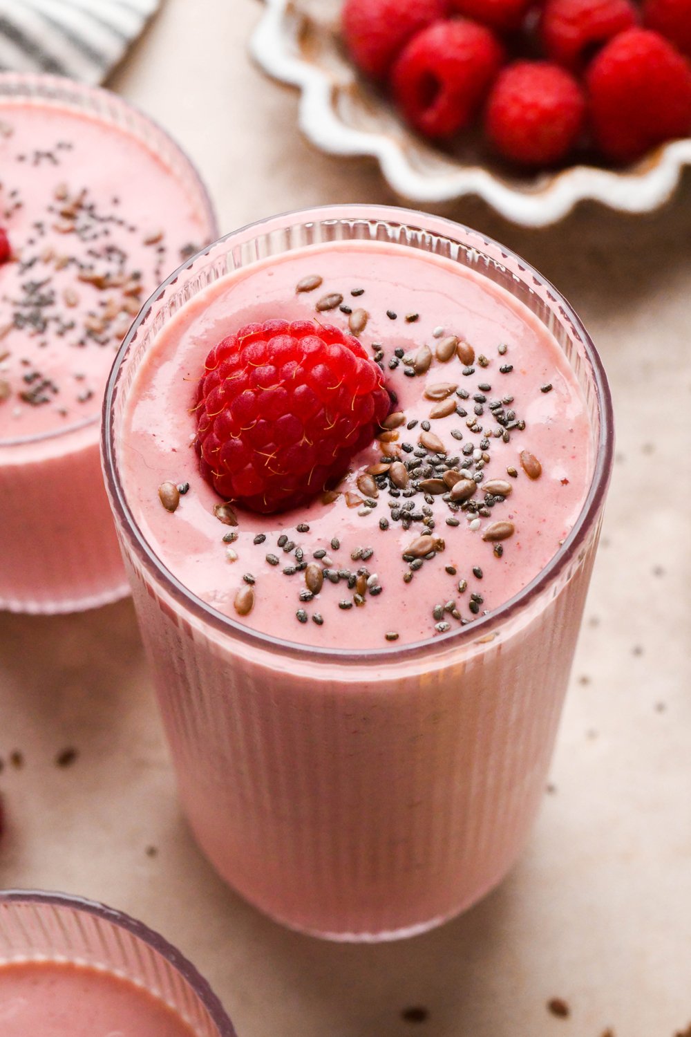 A tall glass of bright pink raspberry mango smoothie next to a smaller glass of smoothie. Both are topped with a single fresh raspberry and a scattering of chia seeds and flax seeds.