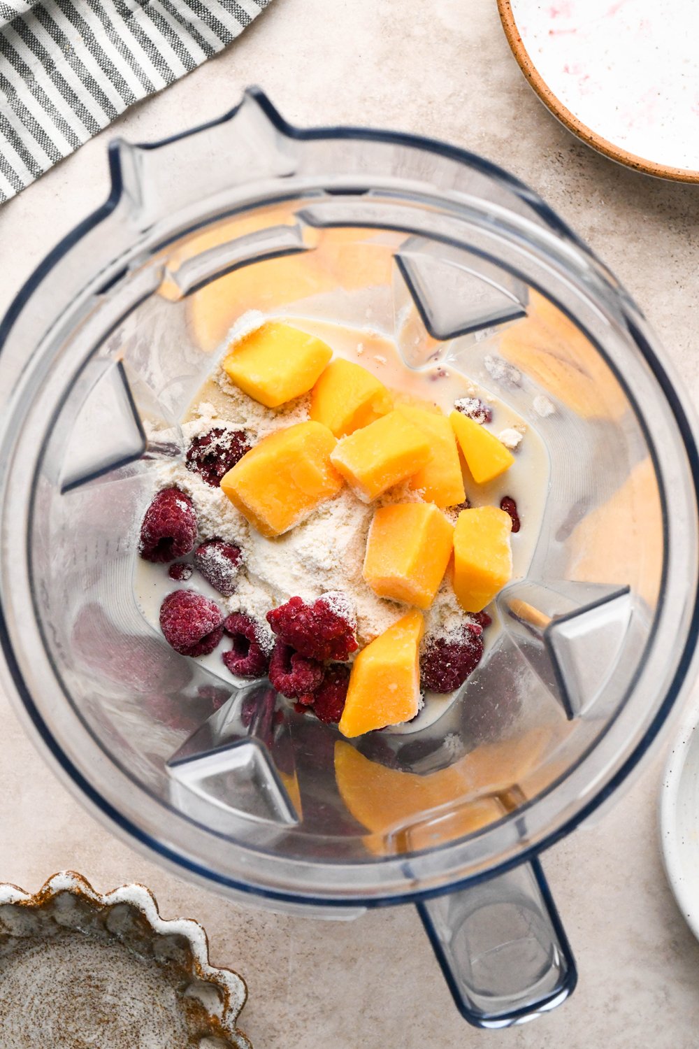 Step-by-step for how to make Raspberry Mango Smoothie with Greek Yogurt: Dairy free milk, yogurt, protein powder, frozen raspberries, and frozen mango in blender container.
