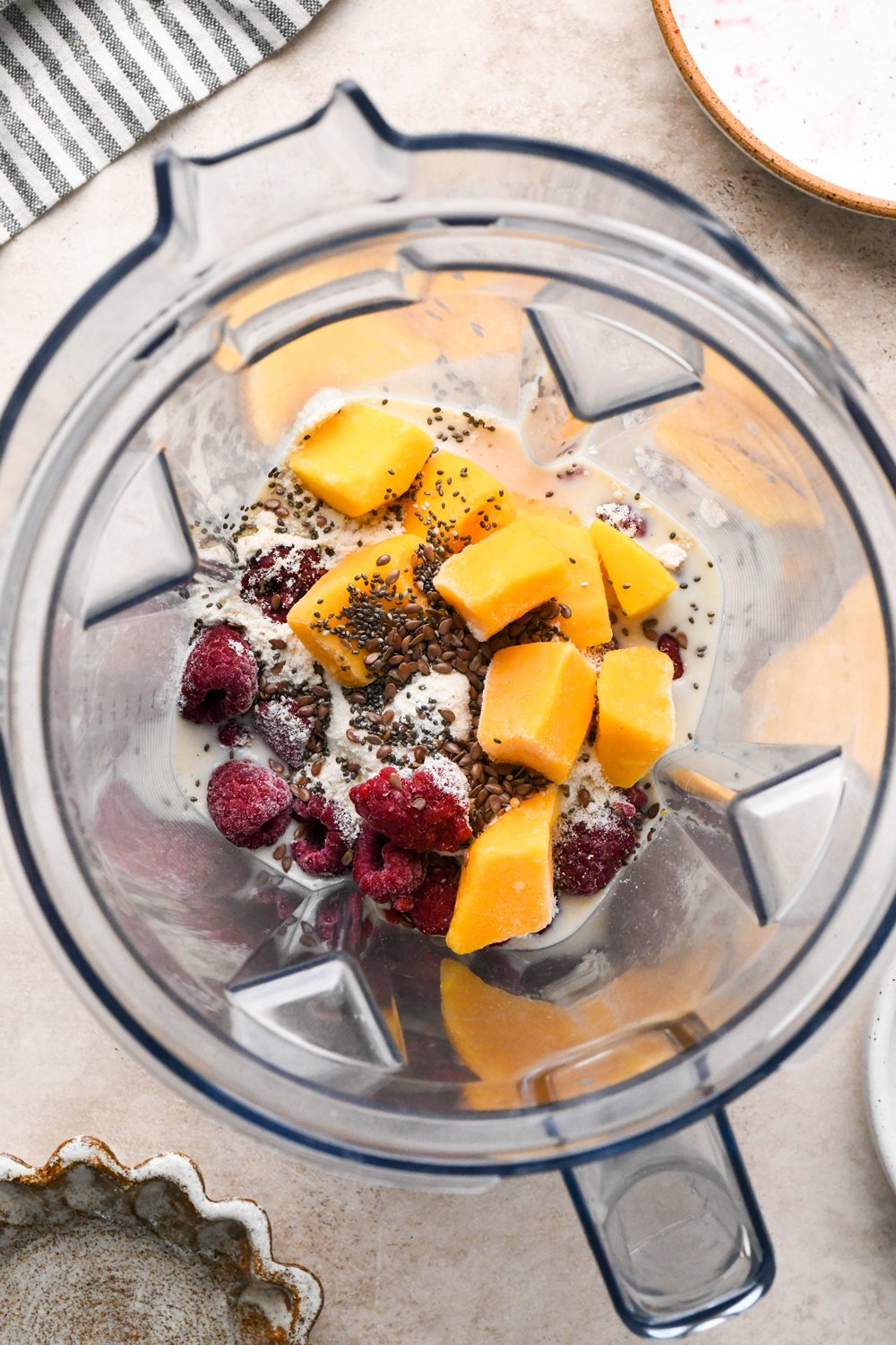 Step-by-step for how to make Raspberry Mango Smoothie with Greek Yogurt: Dairy free milk, yogurt, protein powder, frozen raspberries, frozen mango, chia seeds, and flax seeds in blender container.