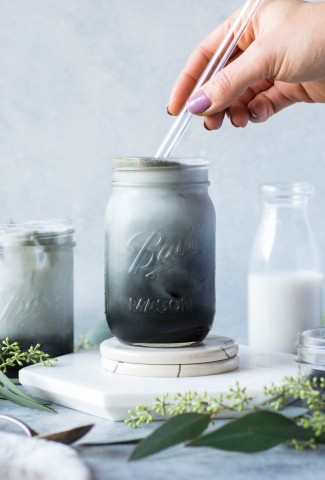 Black and grey toned iced charcoal and almond milk latte in a mason jar, on a grey / blue background with a hand putting a glass straw into the cup.