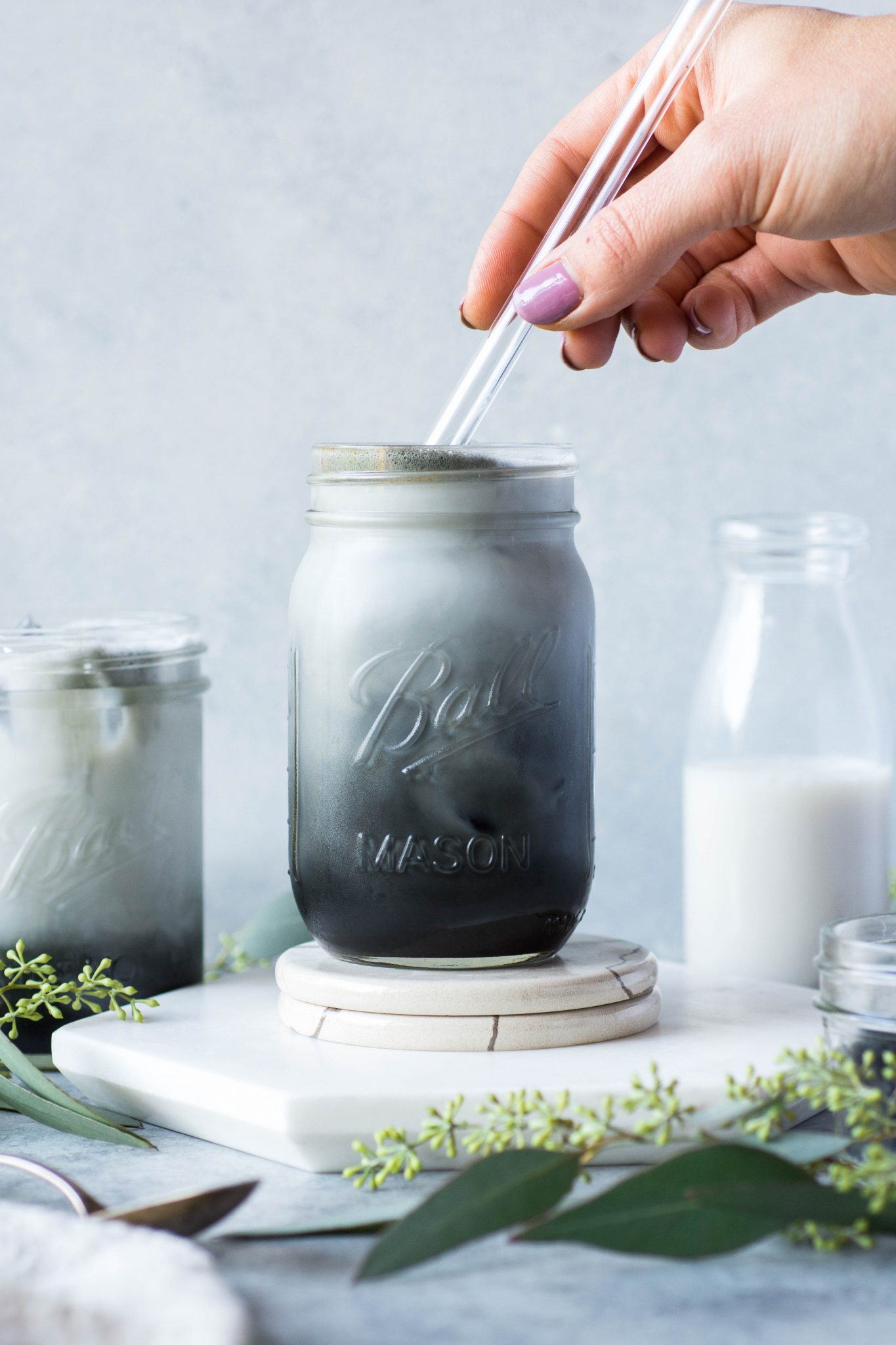 Black and grey toned iced charcoal and almond milk latte in a mason jar, on a grey / blue background with a hand putting a glass straw into the cup.