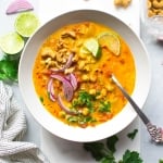 Large white bowl of bright orange chickpea and tomato coconut curry soup. Soup is topped with fresh red onion slices, chopped herbs, curry spiced cashews, and lime wedges. On a white background next to scattered herbs, lime wedges, a soft striped dish towel, and cashew bits.