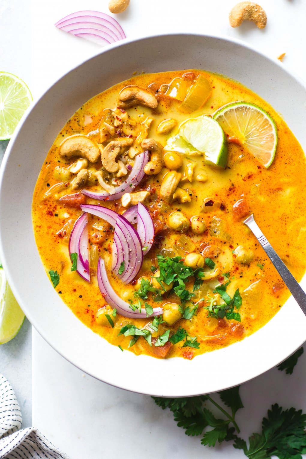 Large white bowl of bright orange chickpea and tomato coconut curry soup. Soup is topped with fresh red onion slices, chopped herbs, curry spiced cashews, and lime wedges. On a white background next to scattered herbs, lime wedges, and cashew bits.