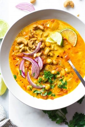 Close up shot of a large white bowl of bright orange chickpea and tomato coconut curry soup. Soup is topped with fresh red onion slices, chopped herbs, curry spiced cashews, and lime wedges. On a white background next to scattered herbs, lime wedges, and cashew bits.