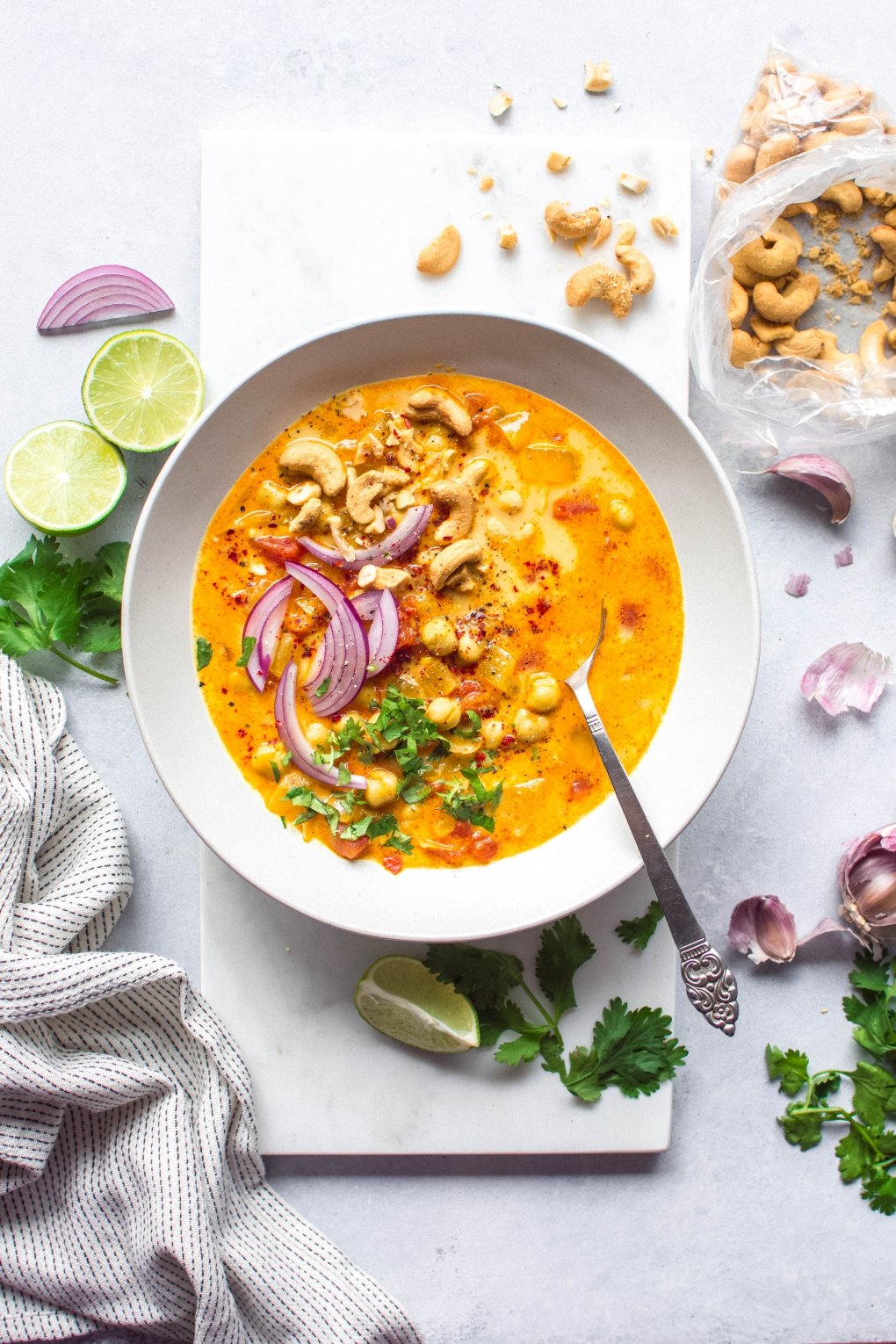 Large white bowl of bright orange chickpea and tomato coconut curry soup. Soup is topped with fresh red onion slices, chopped herbs, curry spiced cashews, and lime wedges. On a white background next to scattered herbs, lime wedges, and cashew bits.