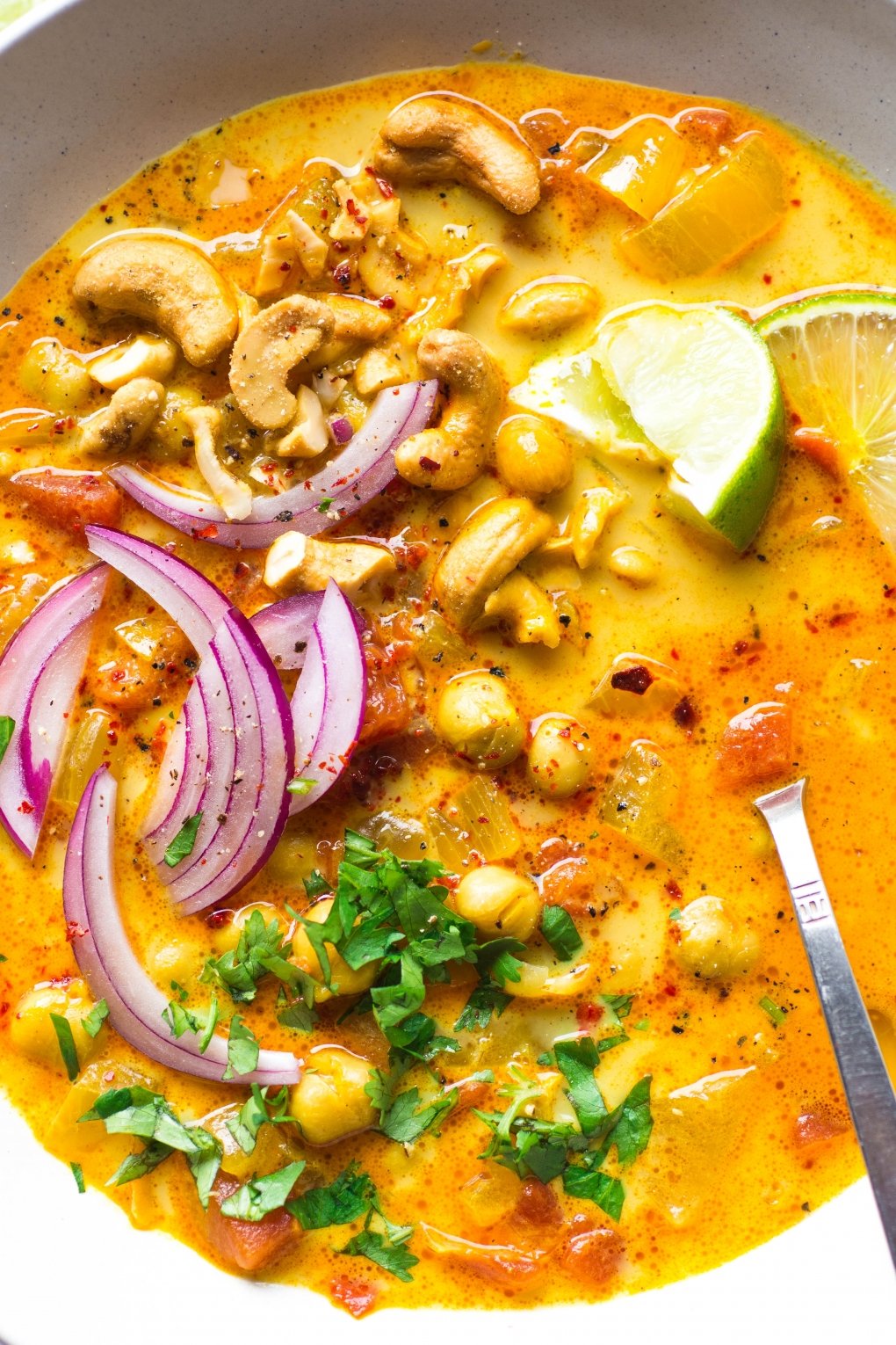 Super close up shot of a large white bowl of bright orange chickpea and tomato coconut curry soup. Soup is topped with fresh red onion slices, chopped herbs, curry spiced cashews, and lime wedges.
