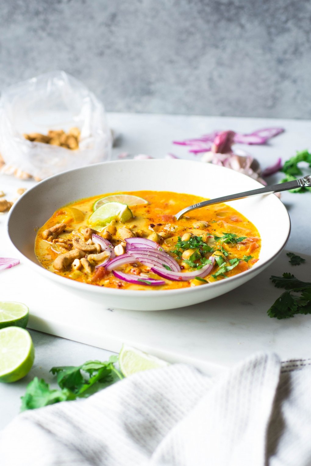 Side angle shot of a large white bowl of bright orange chickpea and tomato coconut curry soup. Soup is topped with fresh red onion slices, chopped herbs, curry spiced cashews, and lime wedges. On a white background next to scattered herbs, lime wedges, and cashew bits.