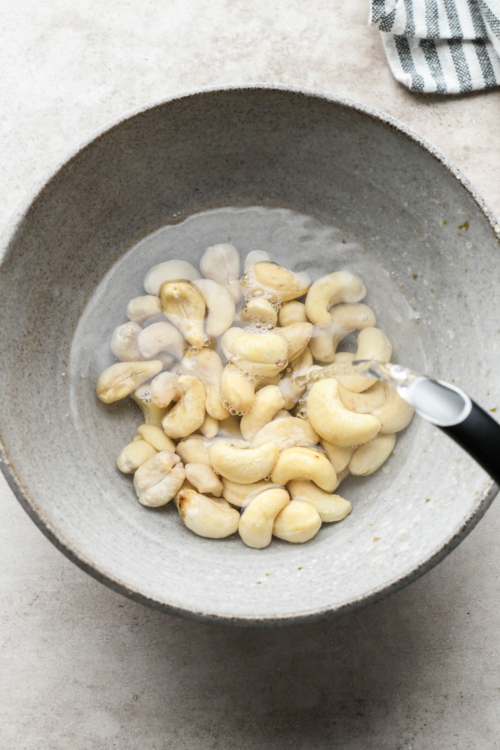 How to make Gluten Free Mushroom Gravy: Pouring boiling water from a tea kettle over raw cashews in a small grey ceramic bowl.