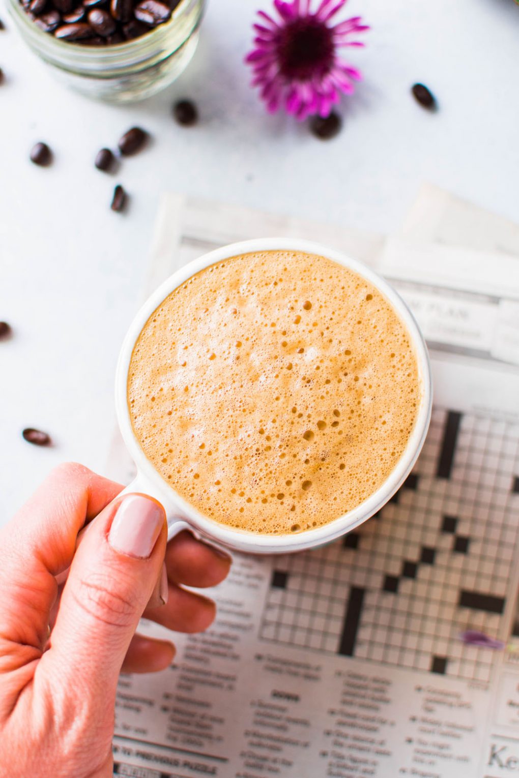 Overhead shot of a hand holding a super creamy cup of bulletproof coffee. On a light background with a newspaper crossword puzzle peeking into the frame. 