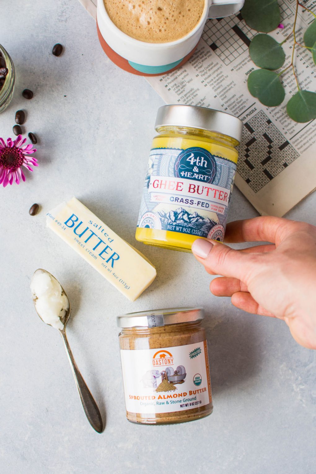 Overhead shot of some fats used for bulletproof coffee. A jar of ghee, a stick of butter, almond butter, and a spoon of coconut oil are all on a light colored background with a newspaper crossword puzzle and a cup of coffee peeking into the frame. 