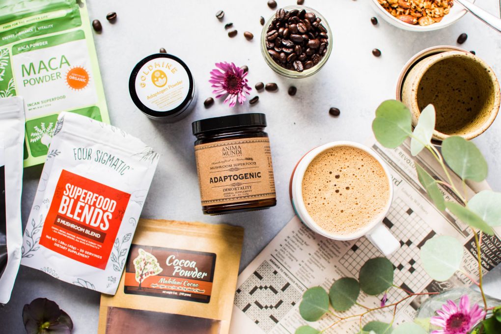 Overhead shot of some adaptogens and flavor additions used for this guide on how to make bulletproof coffee in 3 easy steps. Maca powder, mushroom blend, cacao powder, and a few empty ceramic mugs all on a light colored background with a newspaper crossword puzzle and a cup of bulletproof coffee peeking into the frame. 