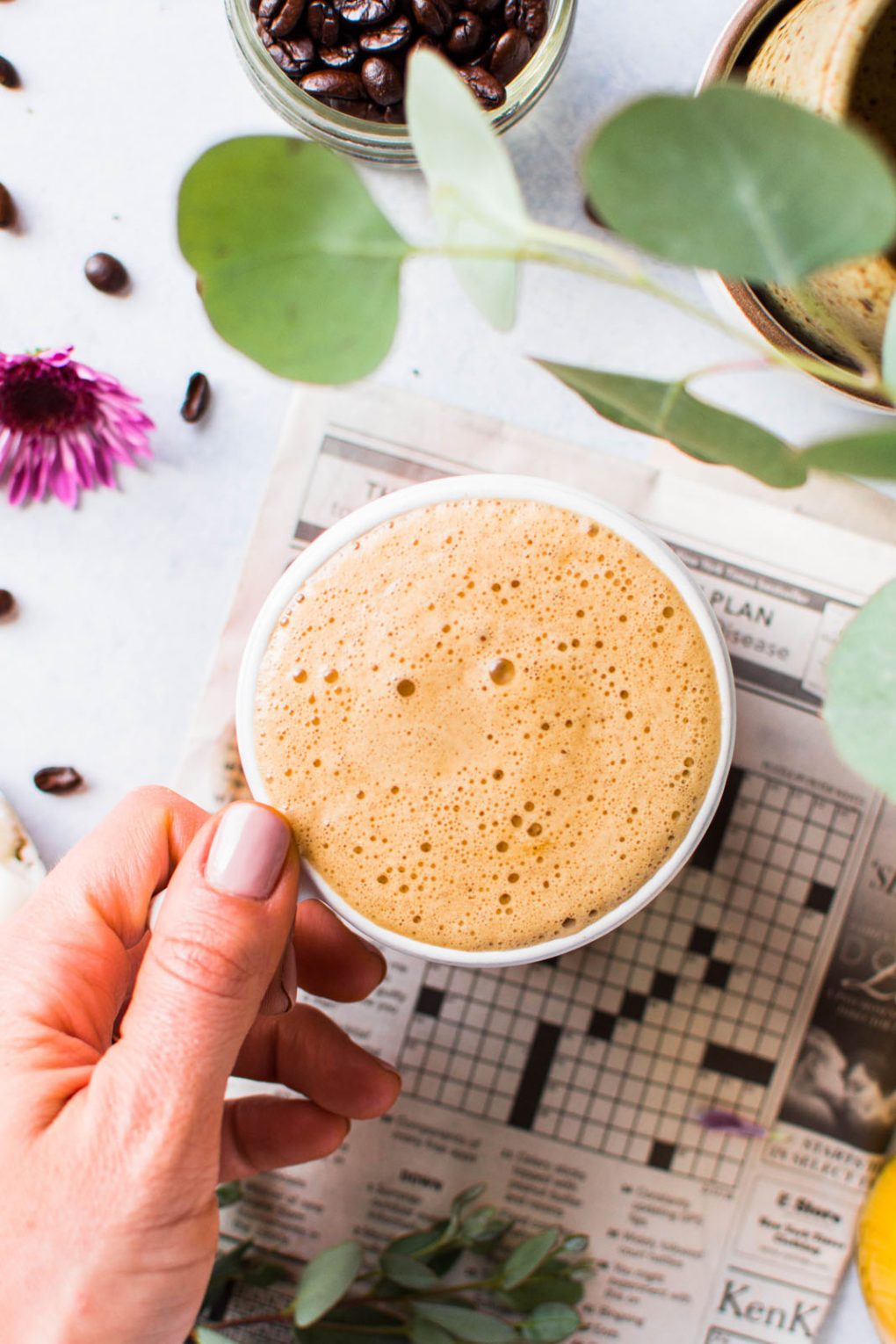 Overhead shot of a hand holding a super creamy cup of bulletproof coffee for a how to make bulletproof coffee in 3 easy steps guide. On a light background with a newspaper crossword puzzle peeking into the frame. 