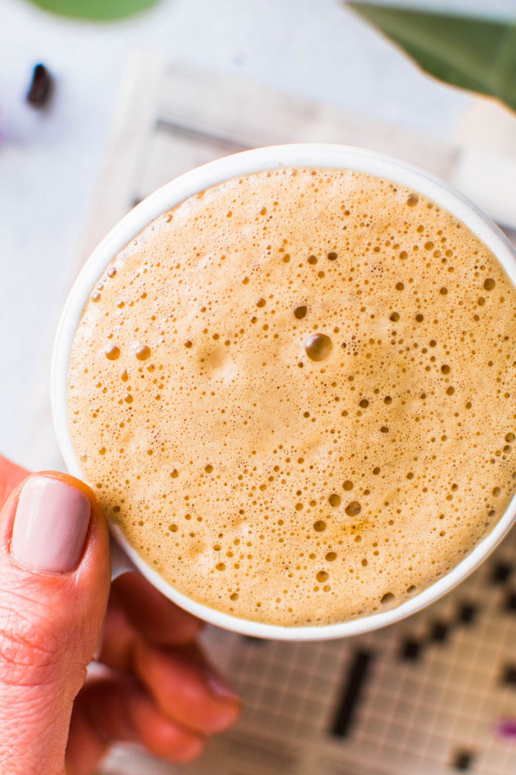 Super close up overhead shot of a hand holding a super creamy cup of bulletproof coffee. On a light background with a newspaper crossword puzzle peeking into the frame. 