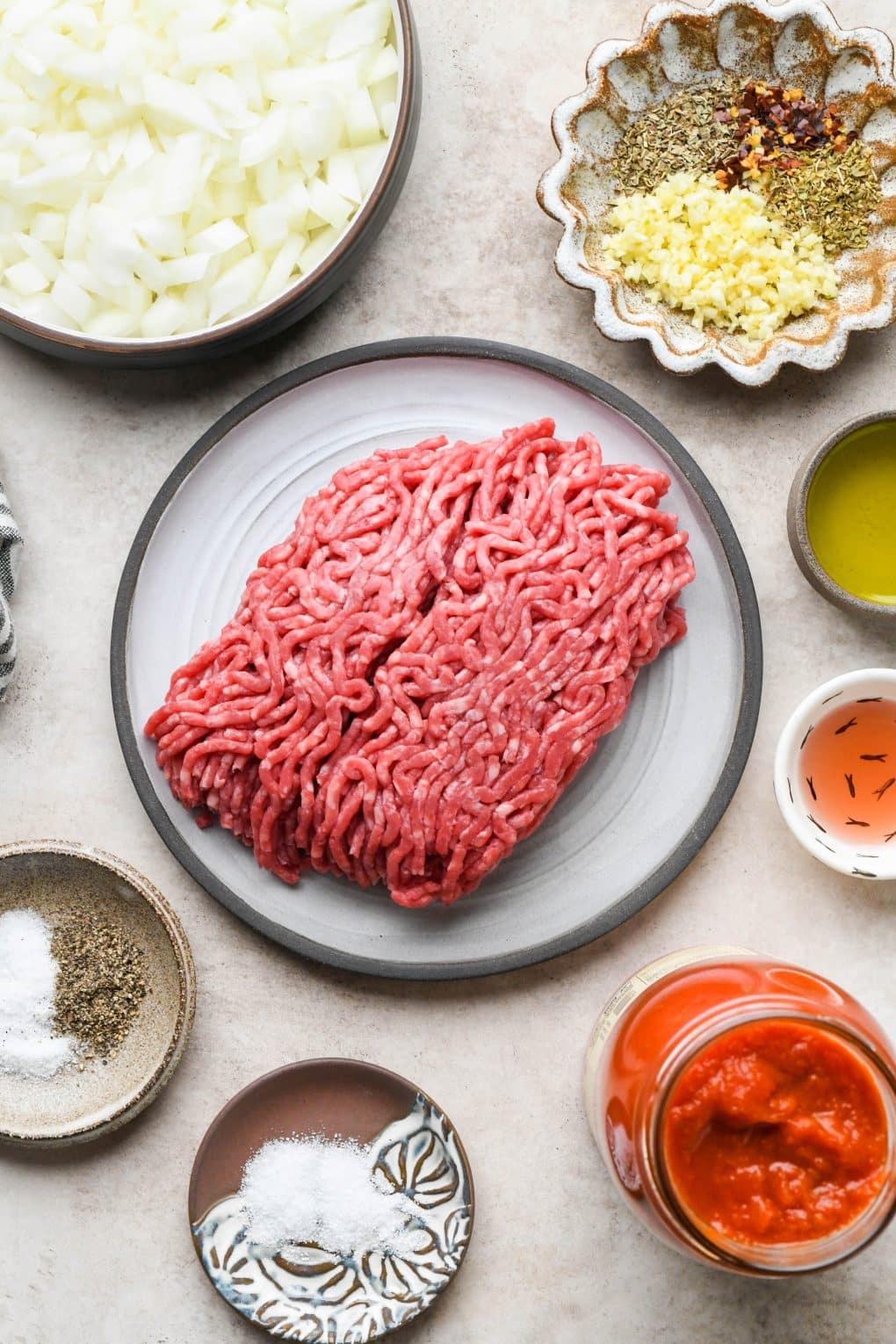Ingredients on various ceramics for marinara sauce with ground beef on a light brown colored background.