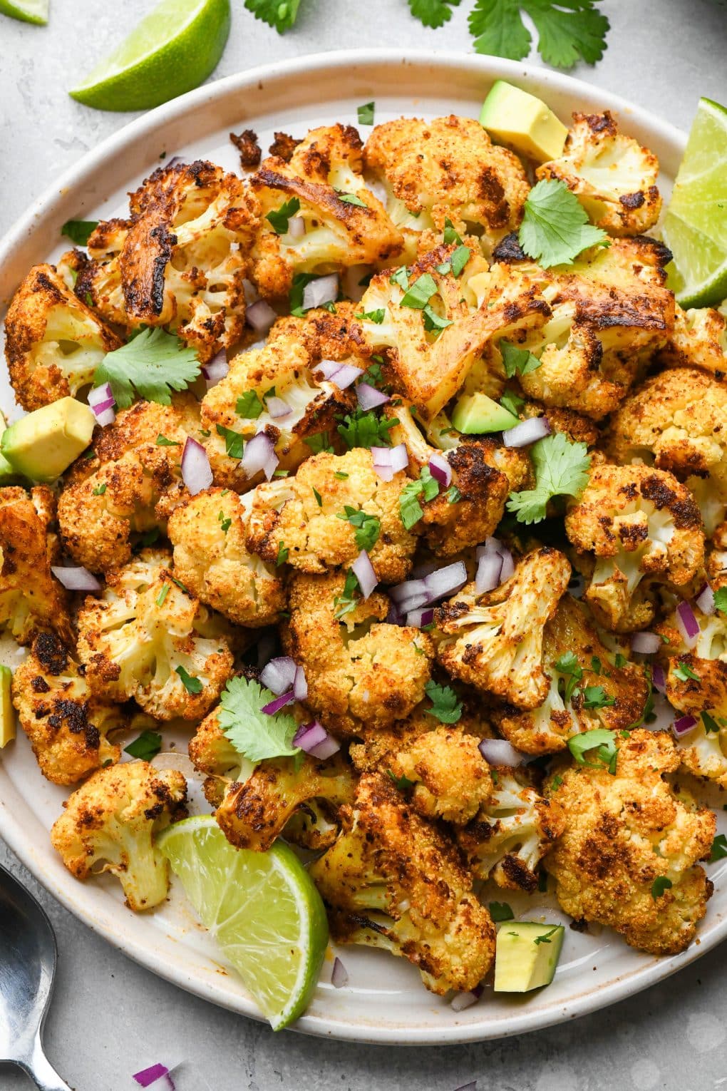 A large plate of Mexican spiced roasted cauliflower, garnished with cilantro, avocado, lime wedges, and finely diced red onion.