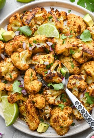 A large plate of Mexican spiced roasted cauliflower with a spoon angled onto the plate, garnished with cilantro, avocado, lime wedges, and finely diced red onion.
