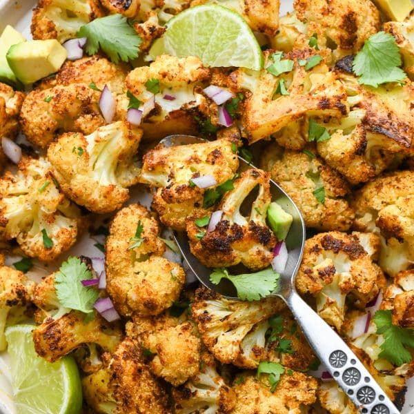 A large plate of Mexican spiced roasted cauliflower with a spoon angled onto the plate, garnished with cilantro, avocado, lime wedges, and finely diced red onion.
