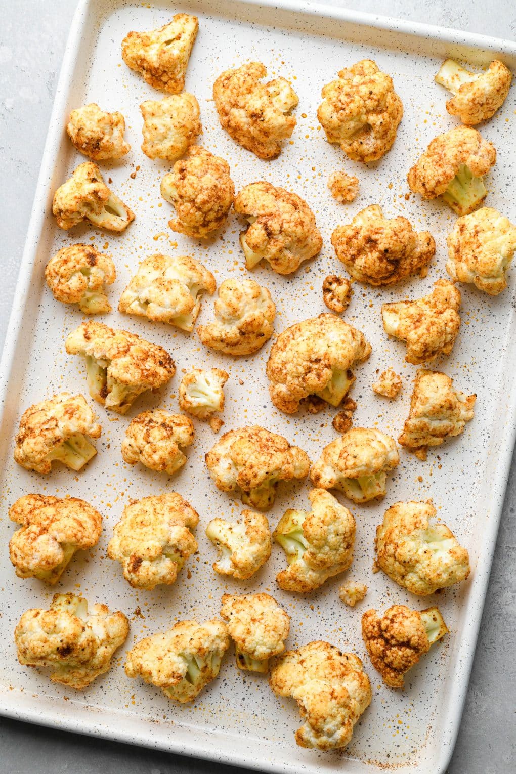 How to make Mexican Roasted Cauliflower: Raw cauliflower florets tossed with oil and spices on a white speckled baking sheet.