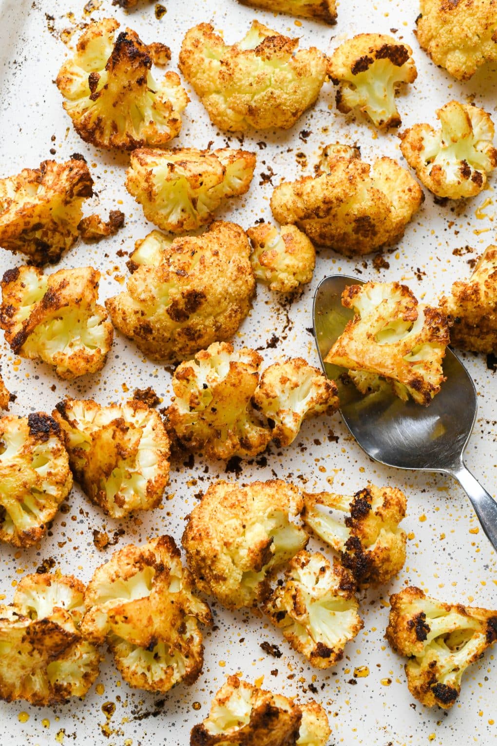 How to make Mexican Roasted Cauliflower: Cooked and crispy cauliflower florets tossed with oil and spices on a white speckled baking sheet, with a spoon holding a few florets.