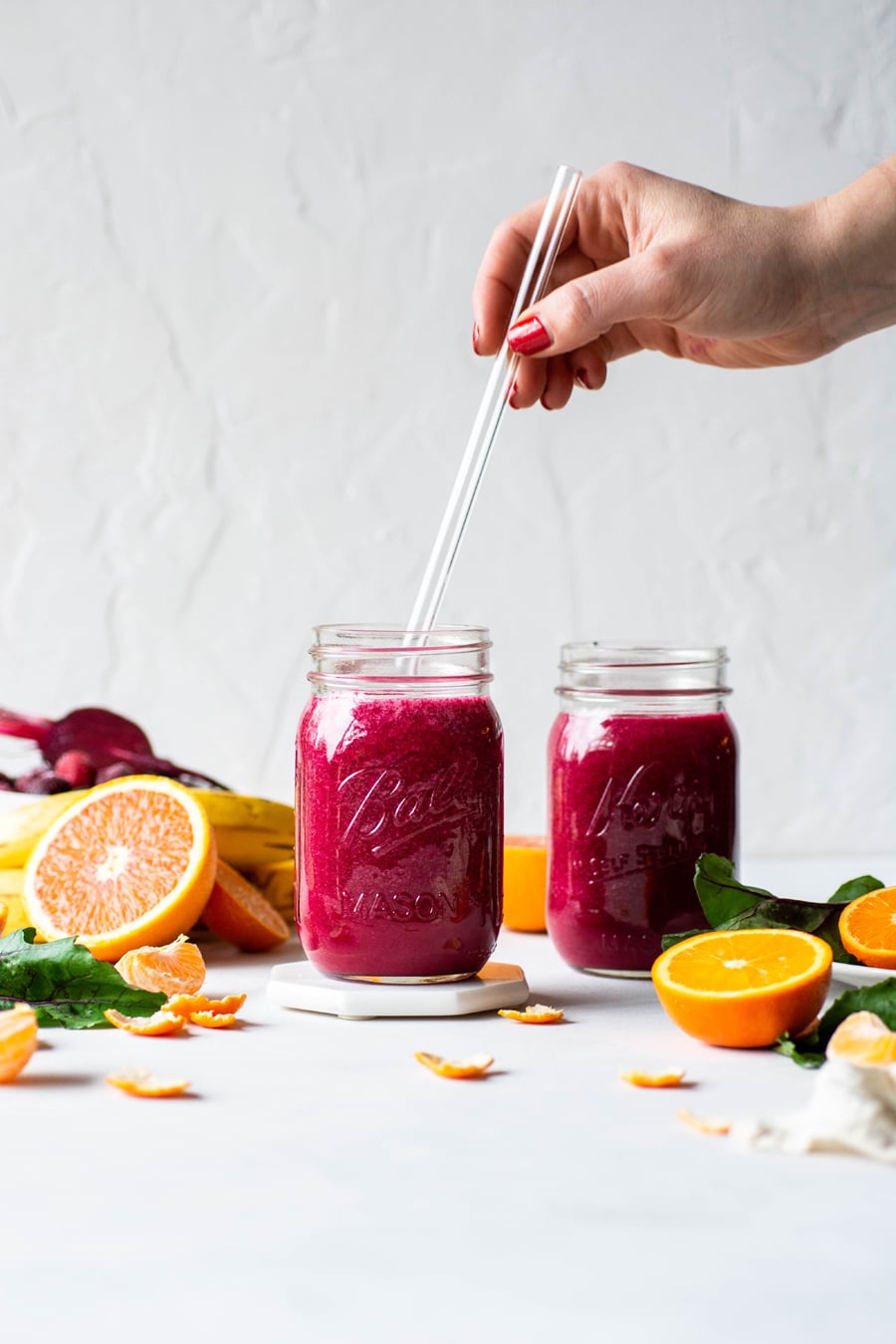 Straight on shot of 2 glass ball jars filled with a vibrant pink / red glowing beet smoothie. On a light background surrounded by fresh cut fruit, with a hand reaching into the frame to place a glass straw into one of the ball jars. 