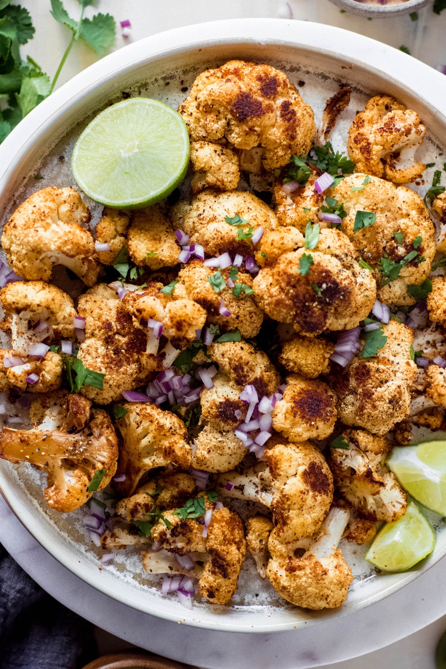 Overhead shot of a plate of mexican roasted cauliflower, topped with chopped red onion, cilantro, and lime wedges.
