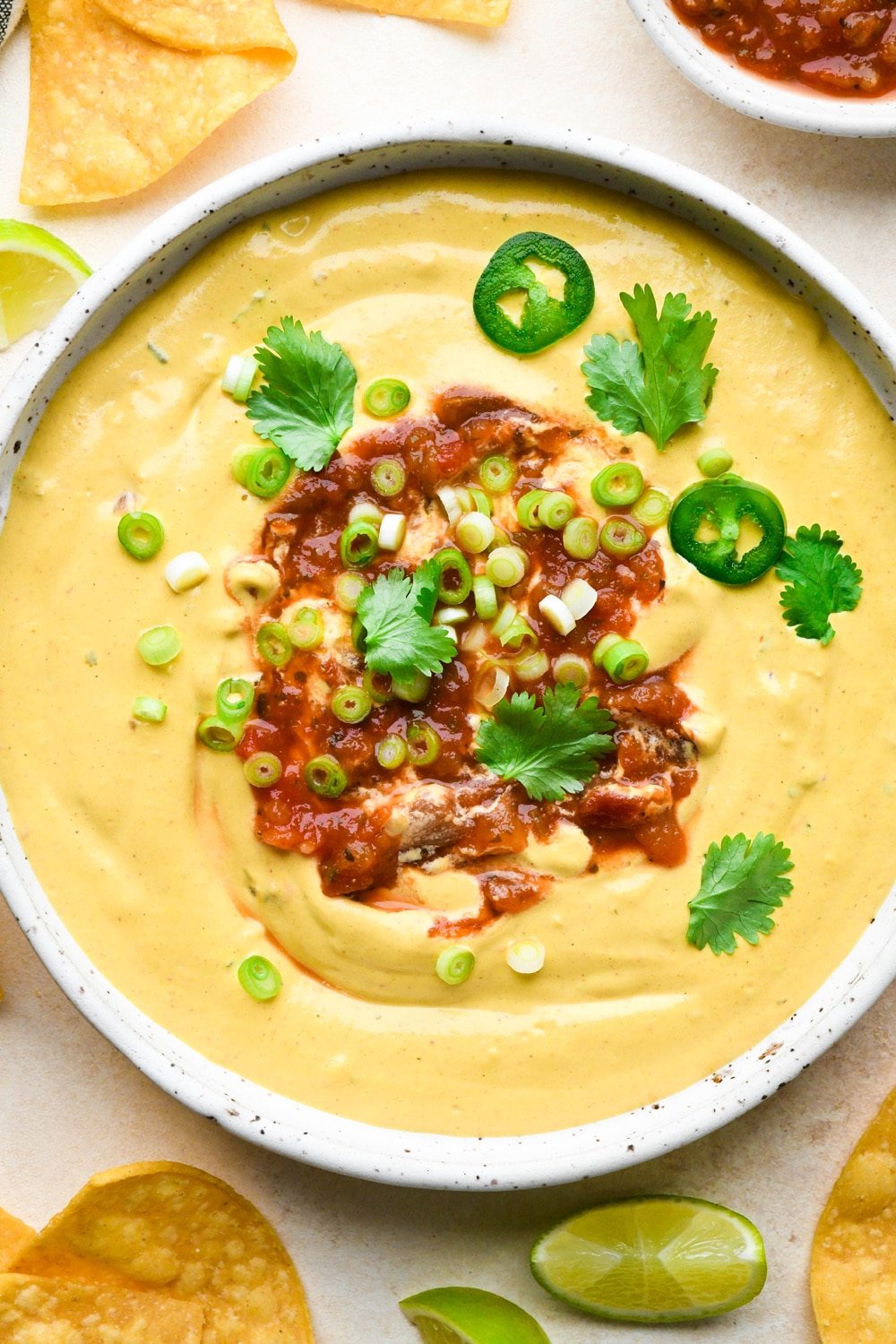 How to make cashew queso: Cashew queso in a shallow ceramic bowl, garnished with a swirl of salsa, green onions, cilantro, and jalapeño slices.